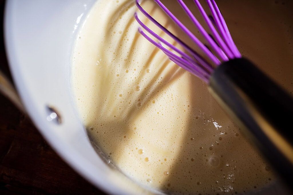 A purple whisk stirs a light, frothy apple creme brulee mixture in a white bowl, casting a shadow across the surface.