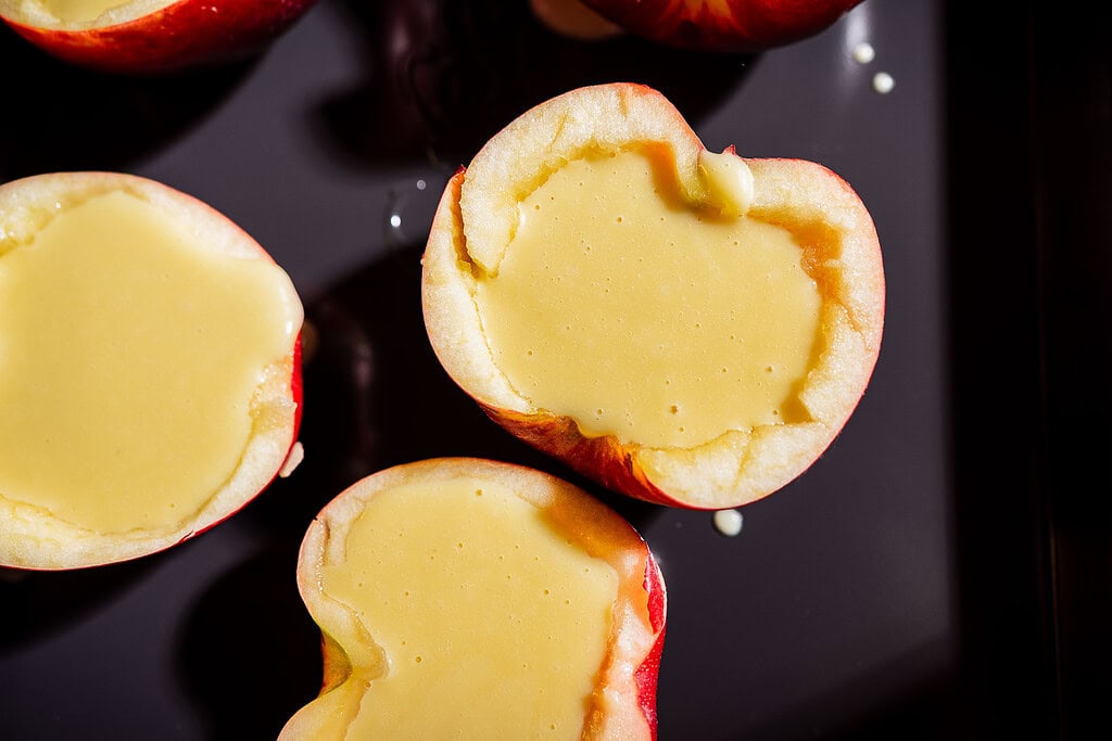 Halved apples with centers filled with a pale yellow apple creme brulee custard, placed on a dark baking sheet.