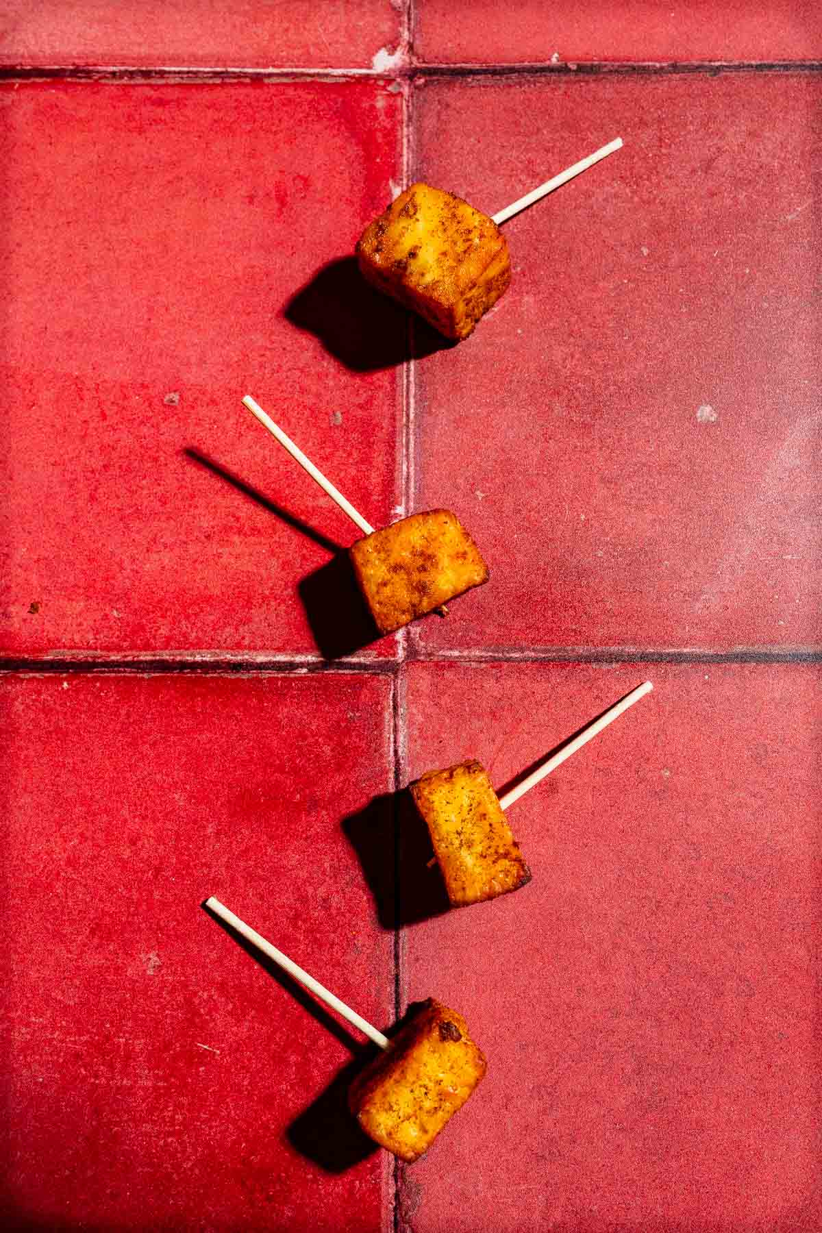 Four air fryer paneer cubes on wooden skewers arranged on a red tiled surface.