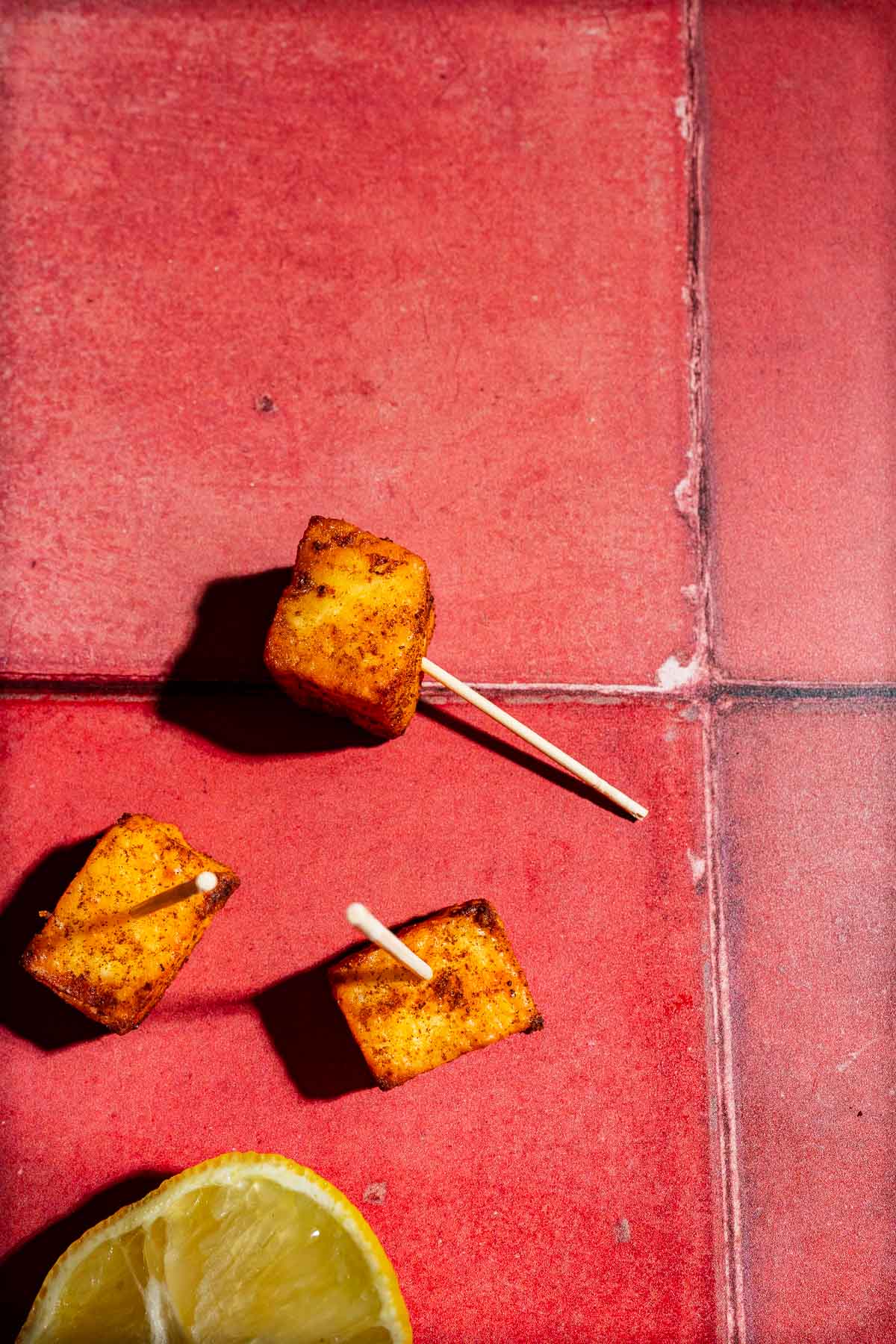 Three cubes of air fryer paneer on toothpicks next to a lemon wedge on a red tiled surface.