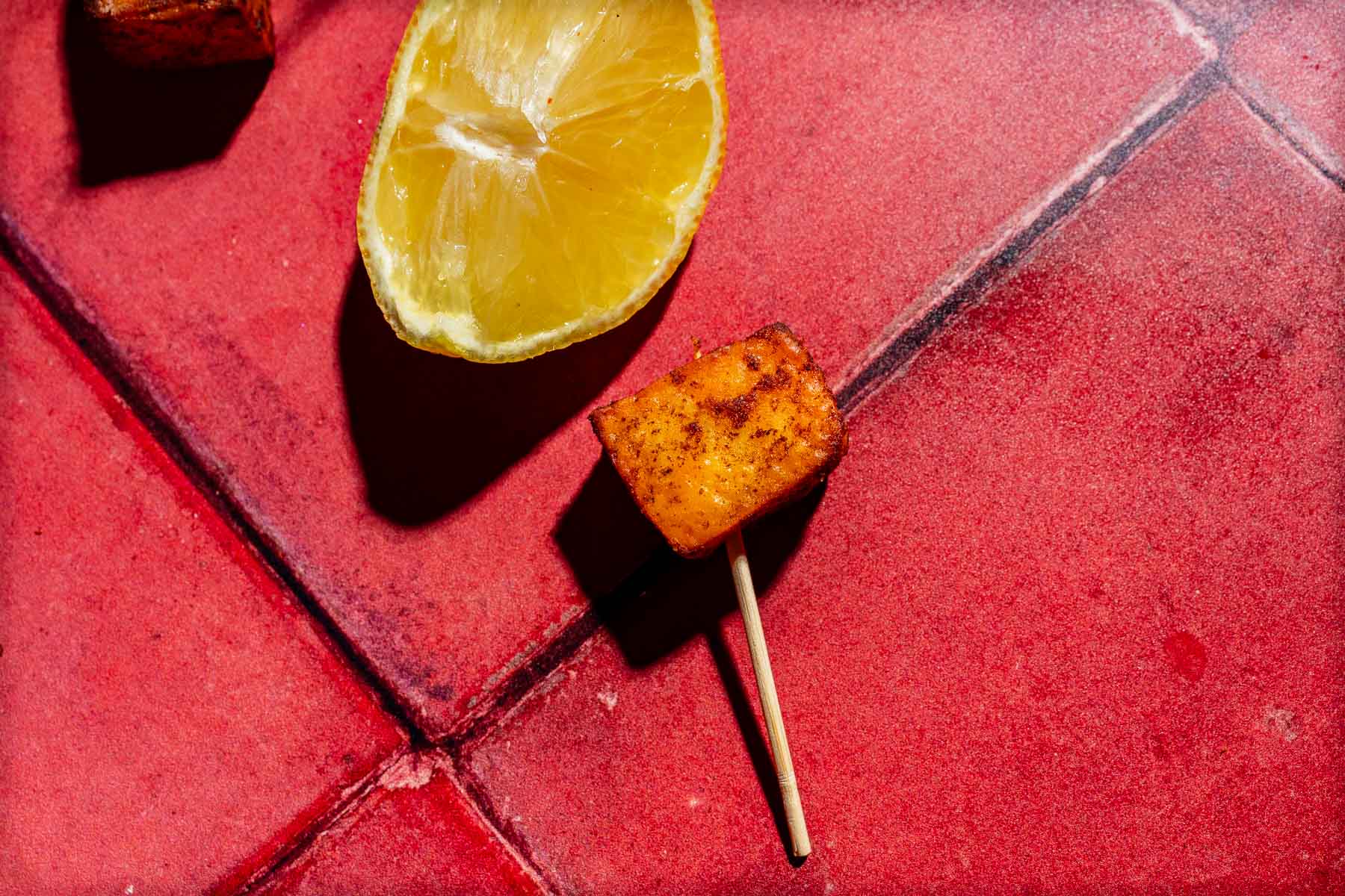 A seasoned cube of air fryer paneer on a wooden pick next to a lemon wedge on a red tiled surface.