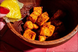 A hand squeezes a lemon wedge over a wooden bowl of seasoned, cubed tofu.
