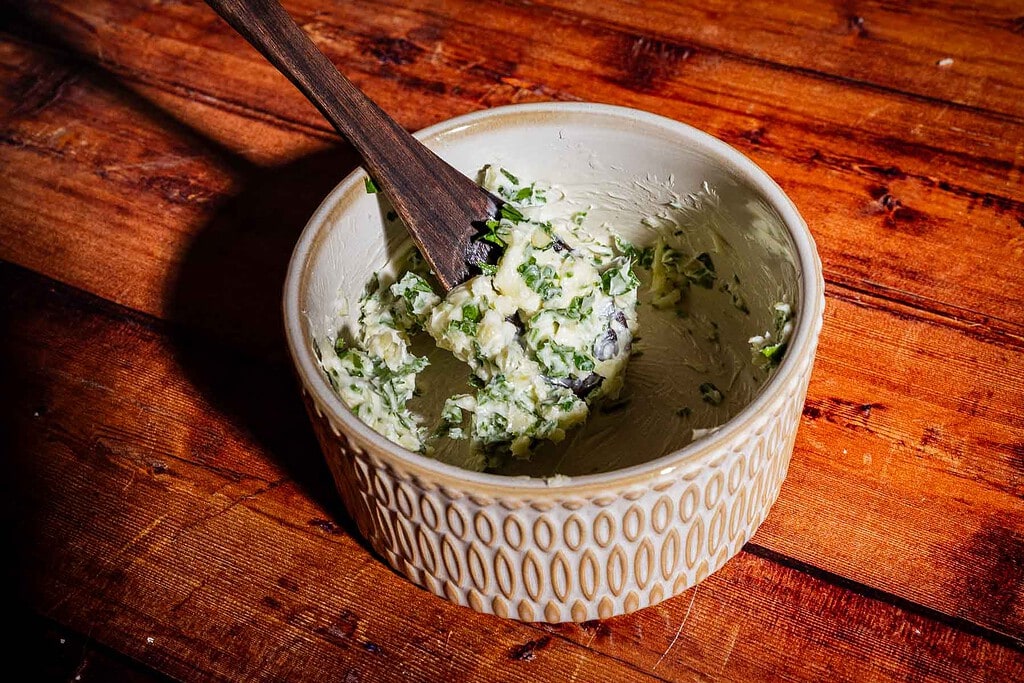 A wooden spoon rests in a ceramic bowl filled with herb butter for puff pastry rolls.