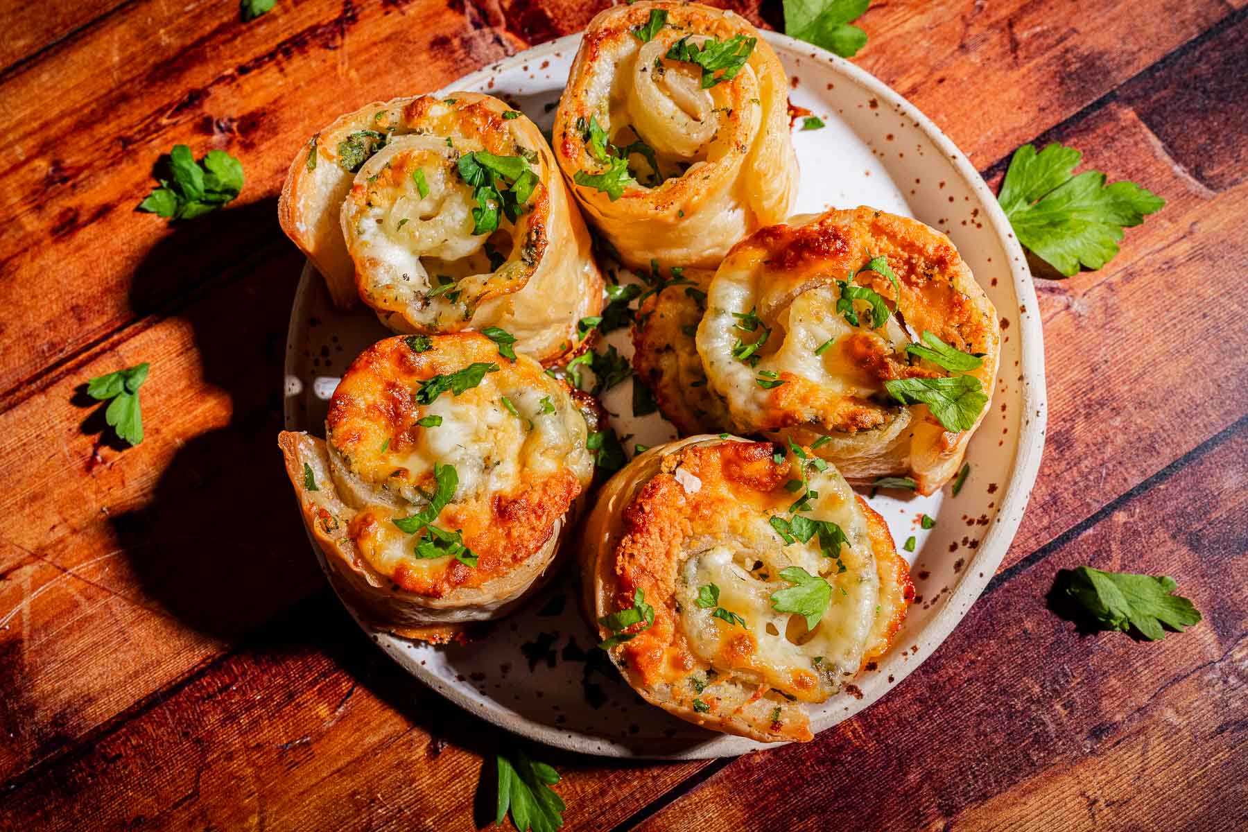 Five savory pastry rolls topped with melted cheese and fresh parsley on a ceramic plate.