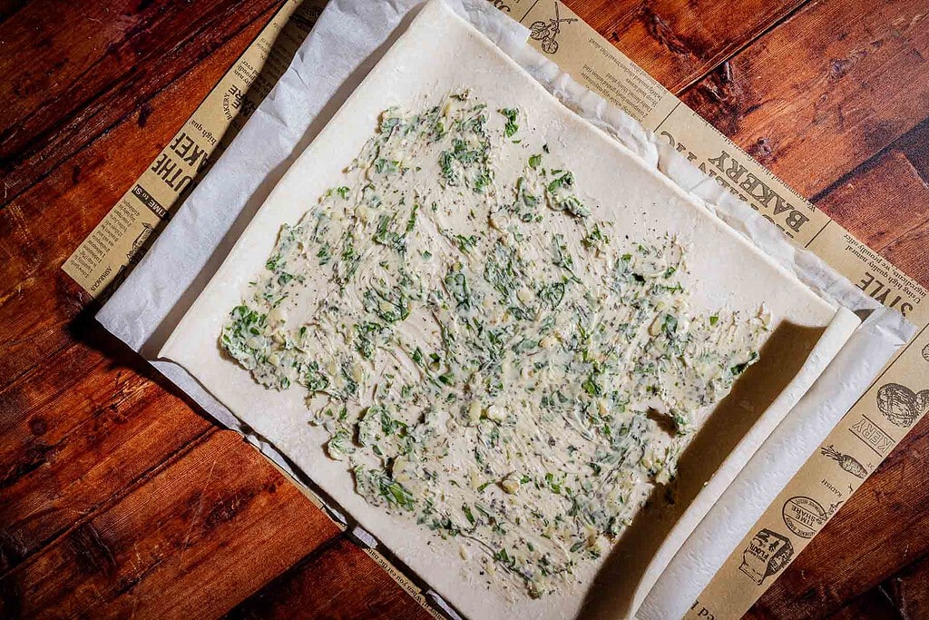 A rectangular sheet of puff pastry dough spread with a mixture of herbs and butter, placed on parchment paper over a wooden surface.