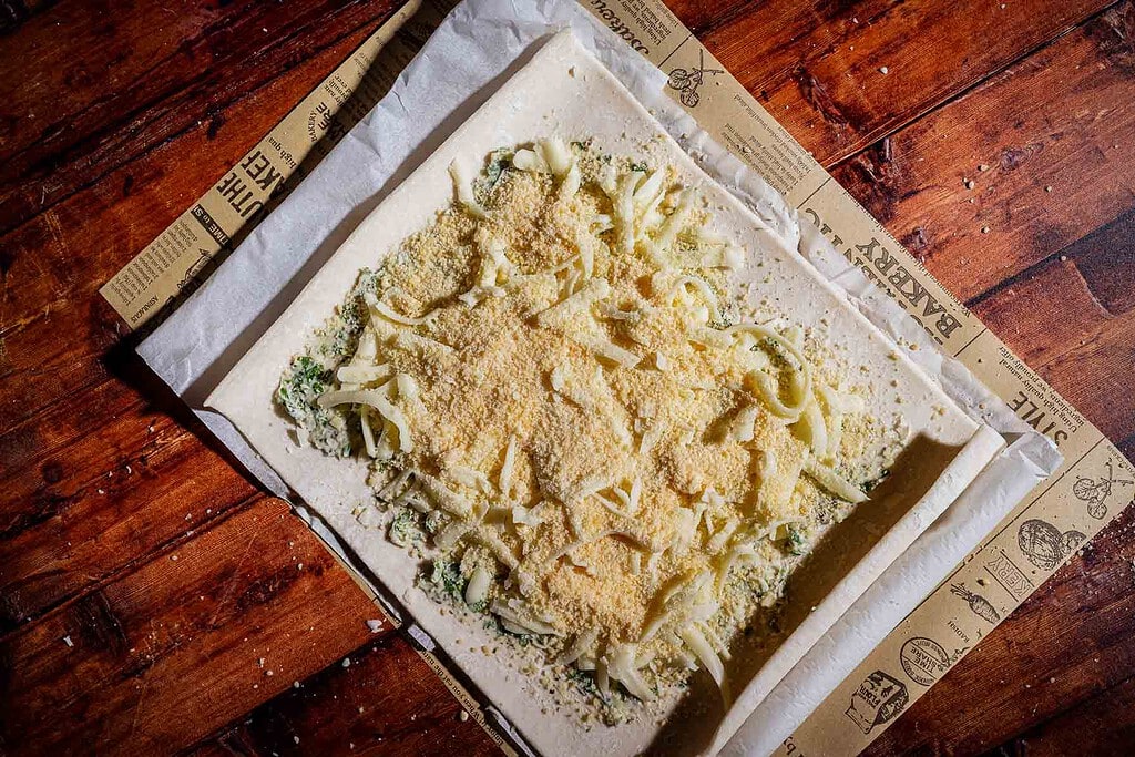 Unbaked puff pastry rolls topped with cheese and herbs on parchment paper.