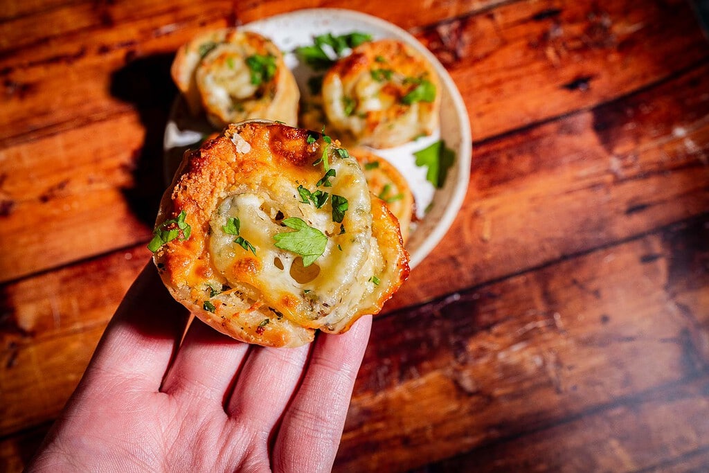 A hand holds a cheesy, herb-topped savory puff pastry roll, with more of the same pastries on a plate in the background.