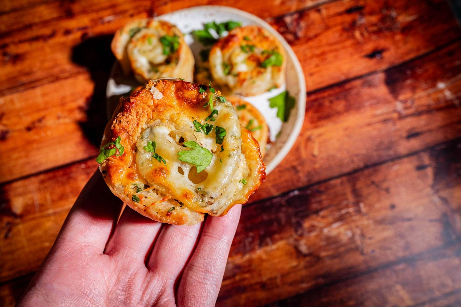 A hand holding a golden-brown savory pastry topped with melted cheese and herbs, with a plate of similar pastries in the background.