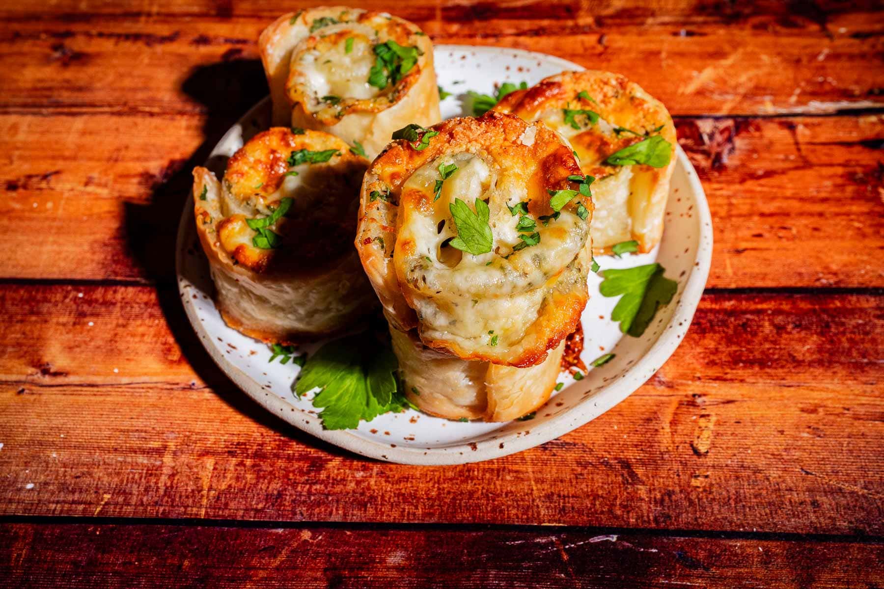 Four savory pastry pinwheels topped with melted cheese and parsley on a speckled plate.