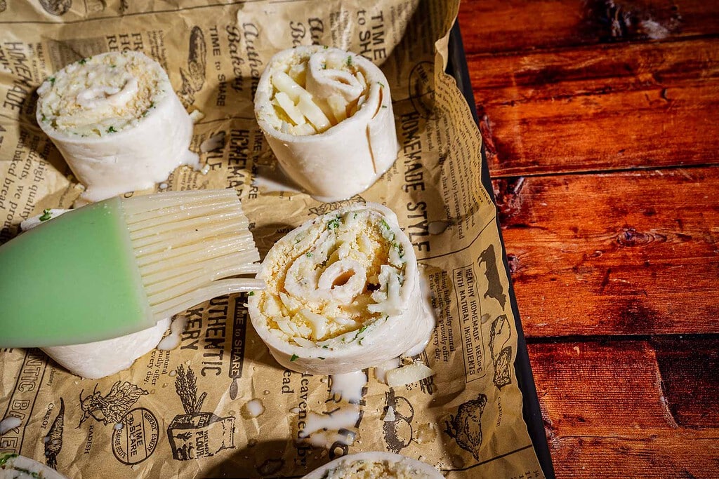 A green silicone pastry brush applies a glaze to three puff pastry rolls filled with cheese and vegetables on printed parchment paper.