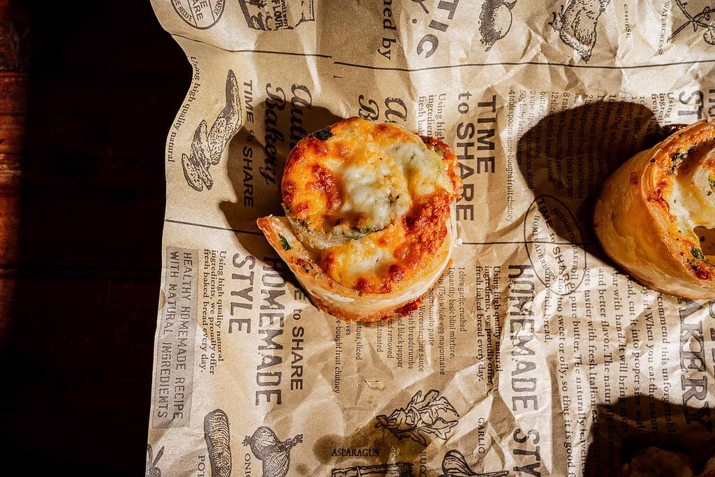 A savory puff pastry roll topped with melted cheese, served on parchment paper printed with bakery-themed text.
