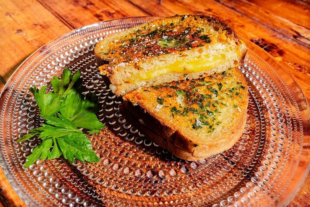 A garlic bread grilled cheese sandwich garnished with herbs on a clear glass plate, served with a sprig of parsley.