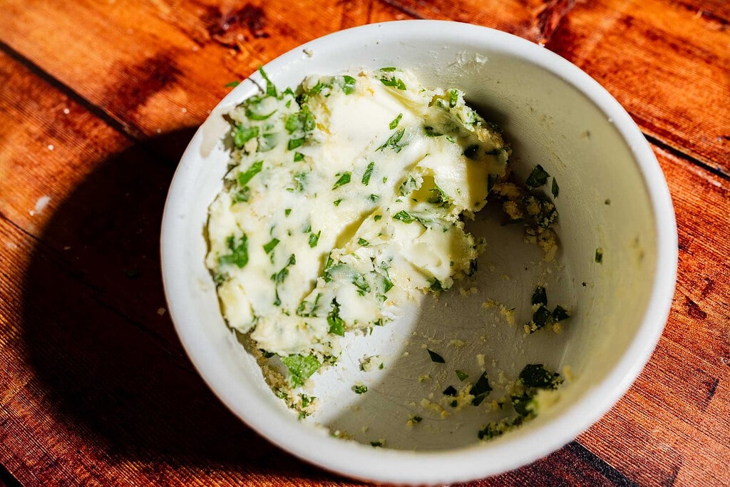A small white bowl containing herb butter mixed with chopped green herbs, perfect for garlic bread.