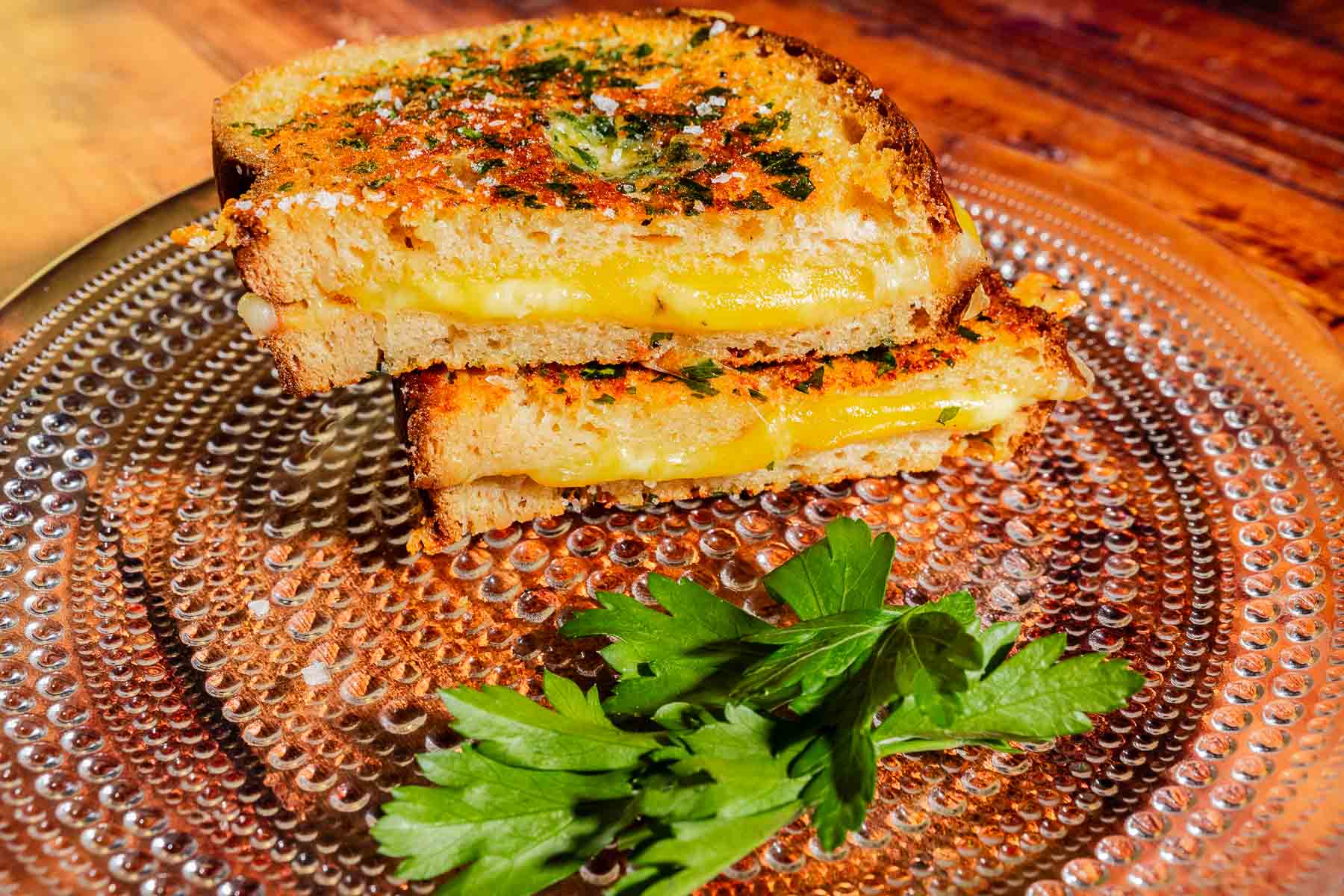 Two halves of a garlic bread grilled cheese sandwich topped with herbs on a textured glass plate with a garnish of parsley.