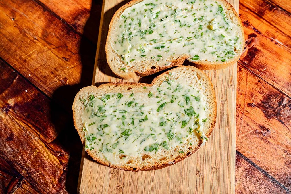 Two slices of garlic bread spread with herb butter on a wooden cutting board.
