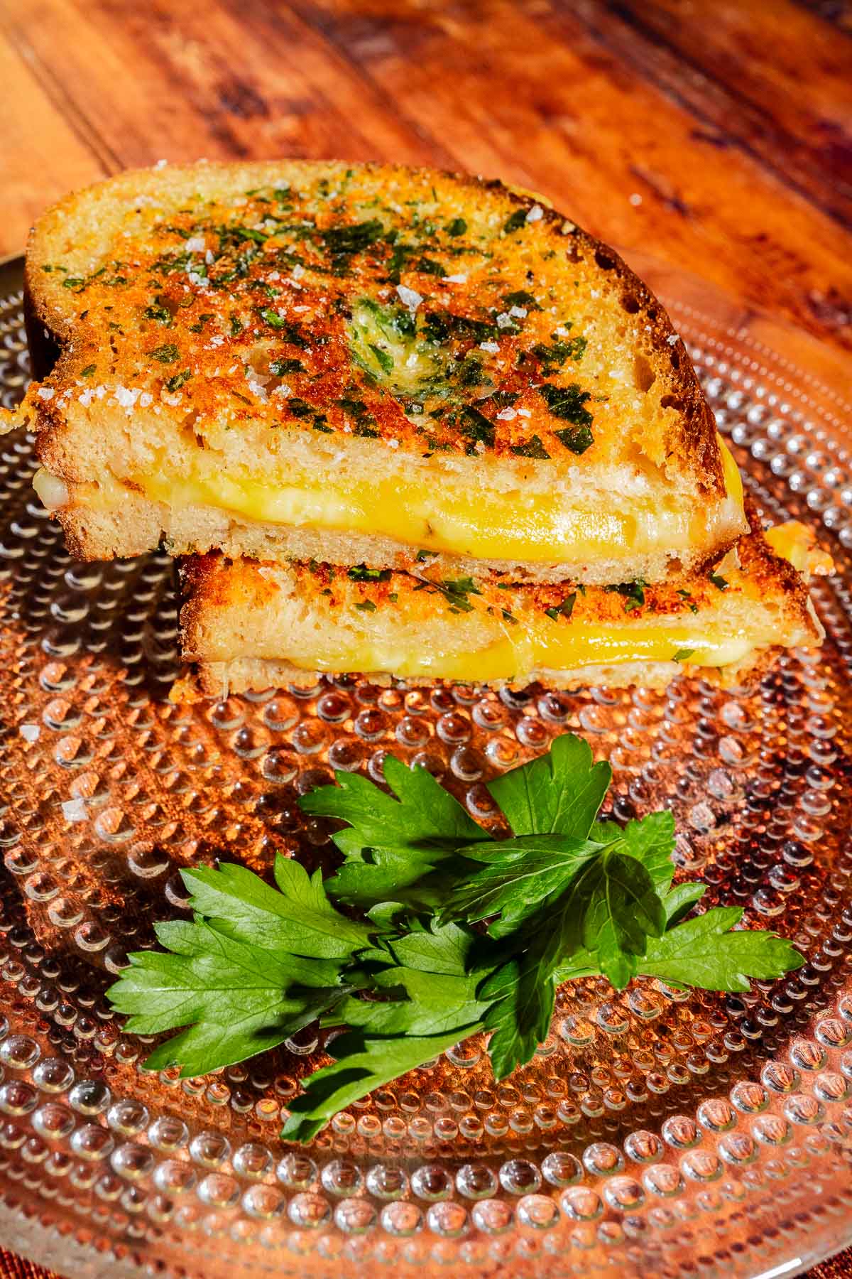 Two halves of a garlic bread grilled cheese sandwich topped with herbs, served with a sprig of parsley on a textured glass plate.