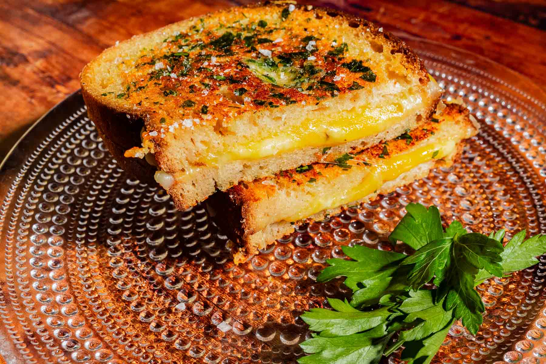A garlic bread grilled cheese sandwich, cut in half and garnished with herbs, on a textured glass plate with a sprig of parsley.
