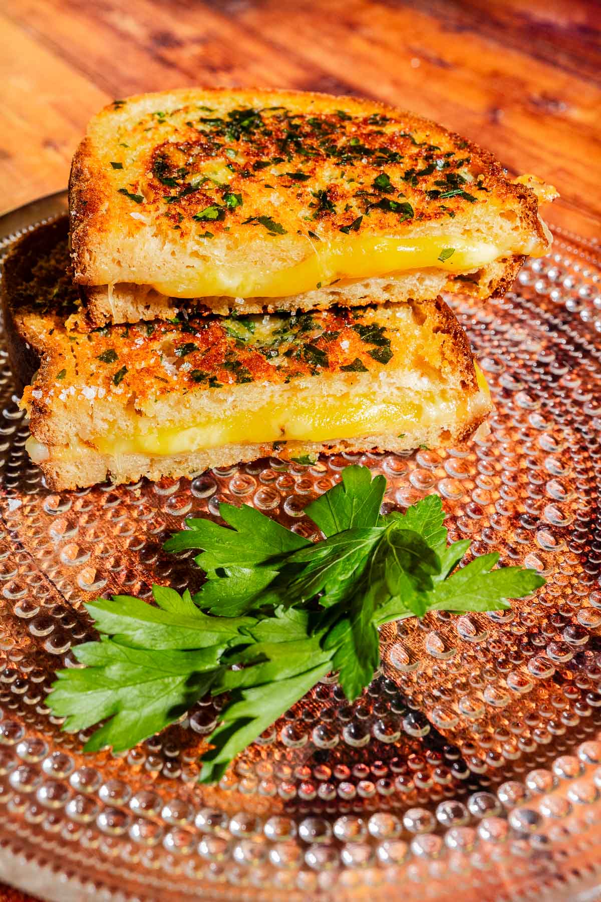Two halves of a garlic bread grilled cheese sandwich topped with herbs on a textured glass plate with a sprig of parsley.