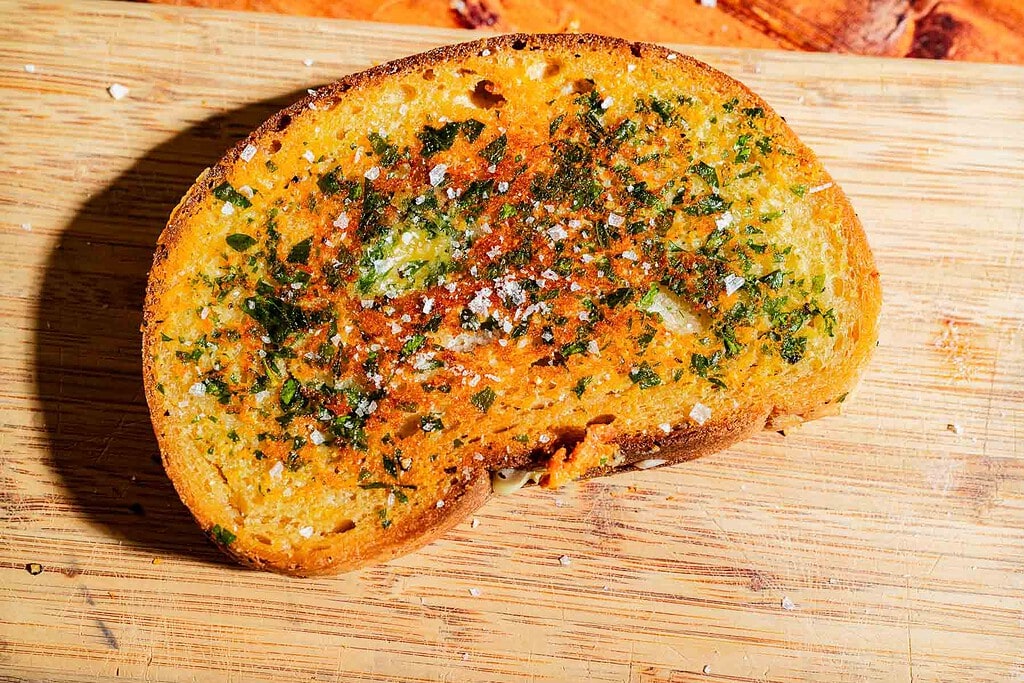 A slice of toasted garlic bread topped with melted butter, herbs, and coarse sea salt on a wooden cutting board.