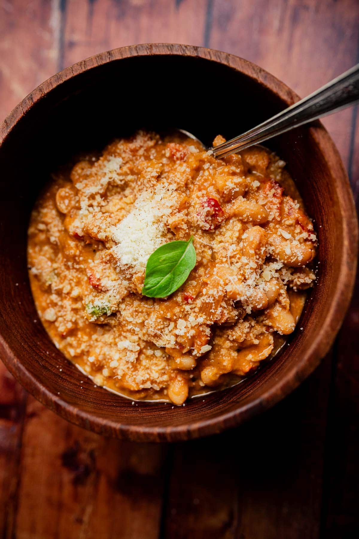 A wooden bowl of marry me chili, a white bean stew topped with grated cheese and a fresh basil leaf, with a metal spoon inside.