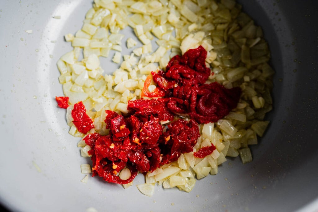 Diced onions and garlic saut&eacute;ing in a pan for a marry me chili, mixed with a dollop of red tomato paste and chopped sun-dried tomatoes.