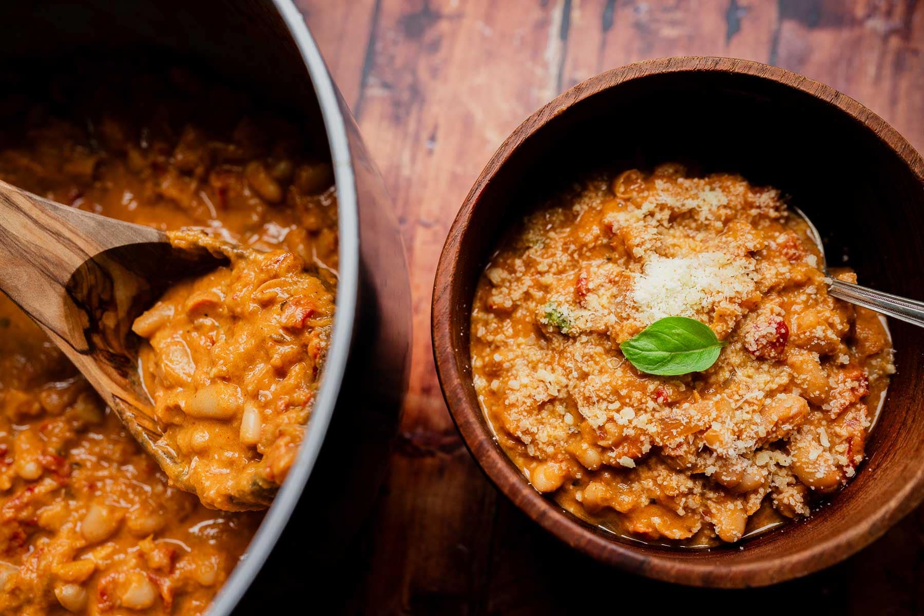 A wooden bowl of creamy marry me chili with white beans, topped with parmesan and basil, next to a pot being served with a wooden spoon.