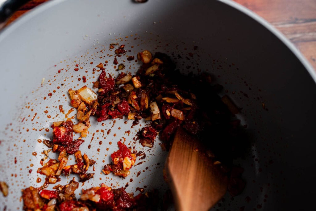 Saut&eacute;ed onions and tomato paste for marry me chili in a grey pot with a wooden spatula.