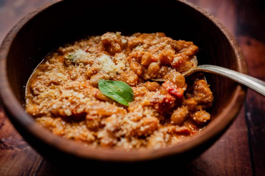 A wooden bowl of marry me chickpea and tomato stew, garnished with grated cheese and a basil leaf, with a spoon resting inside.
