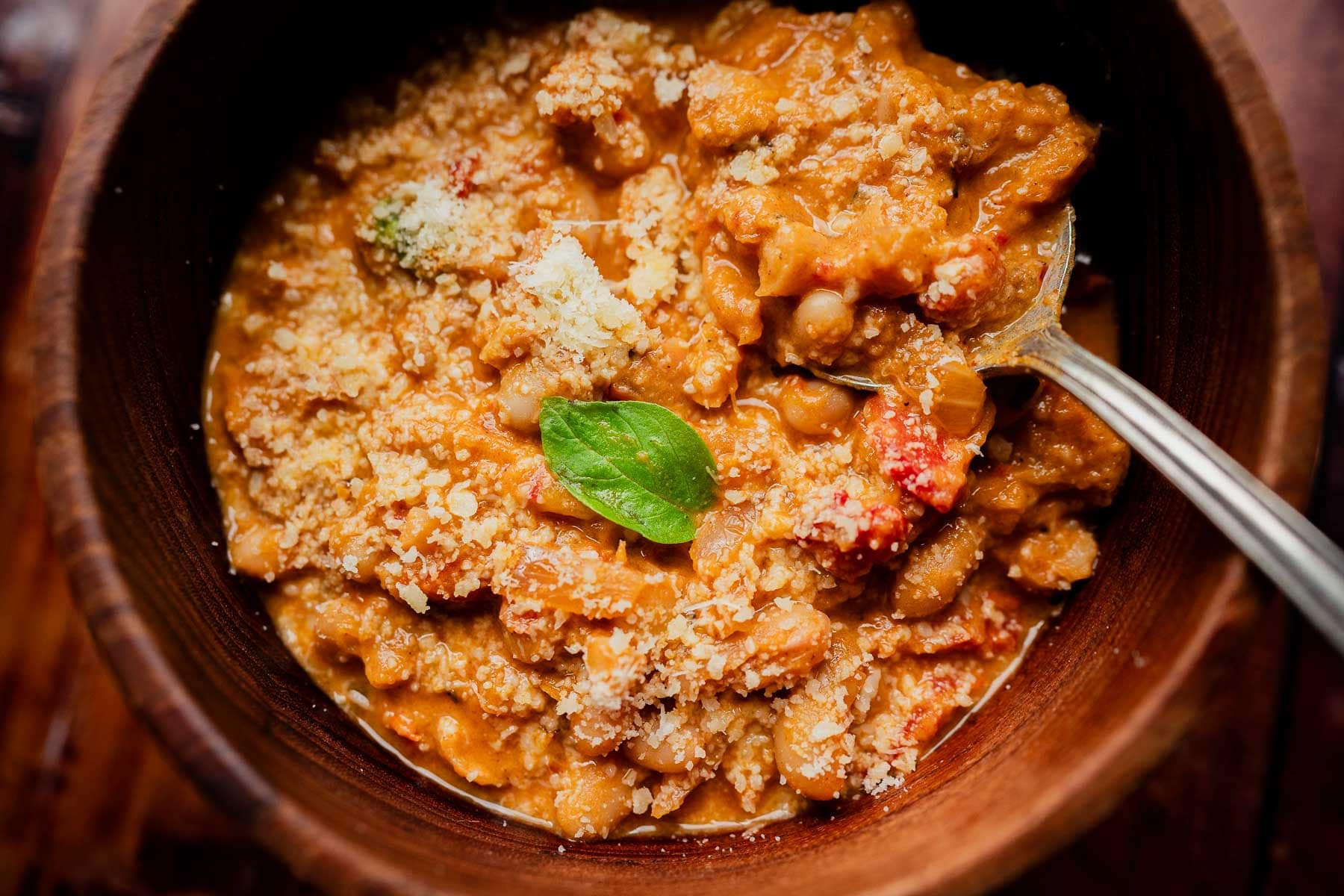 A wooden bowl of marry me chili topped with parmesan and a basil leaf, with a metal spoon resting in the bowl.