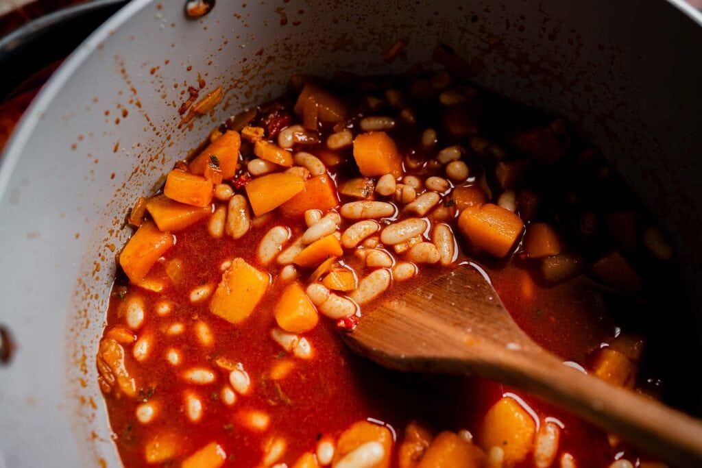 A wooden spoon stirs white beans and cubed squash in a marry me chili red tomato-based sauce inside a cooking pot.