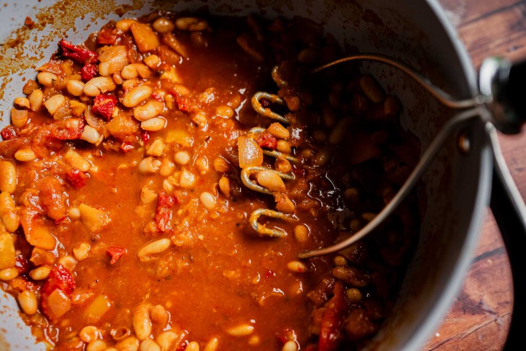 A hand masher pressing into a pot of marry me chili made with stewed white beans and tomatoes.