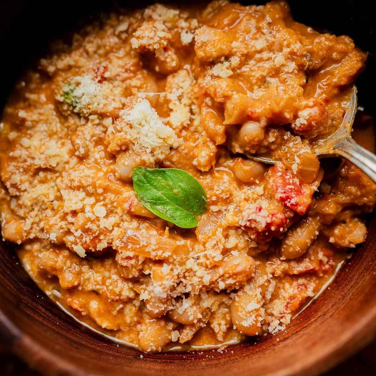 A bowl of marry me chili topped with grated cheese and a fresh basil leaf, with a spoon resting inside.