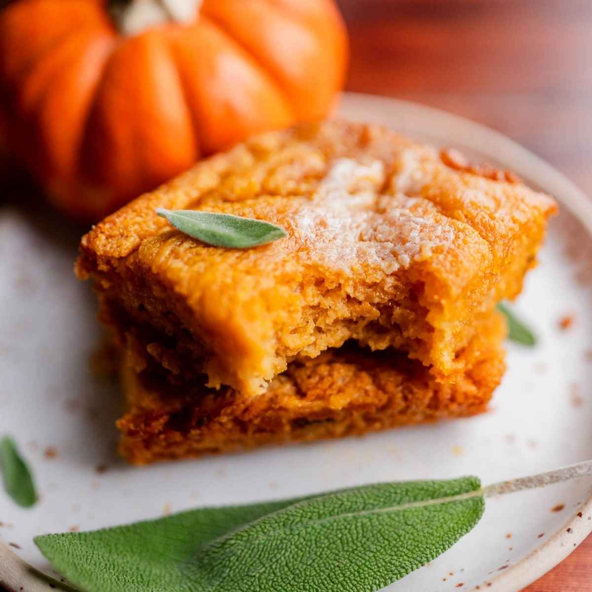 Two stacked squares of pumpkin butter swim biscuits with a bite taken out, garnished with sage leaves, next to a small pumpkin.