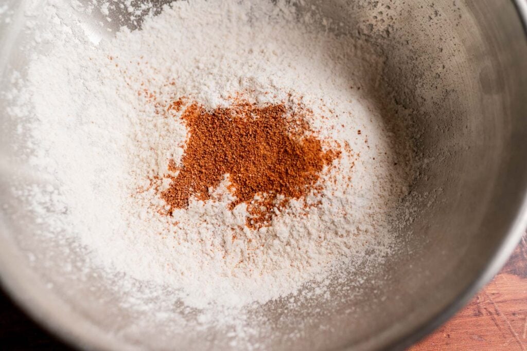 A pile of reddish-brown spice resting on a mound of white flour for pumpkin butter swim biscuits in a metal mixing bowl.