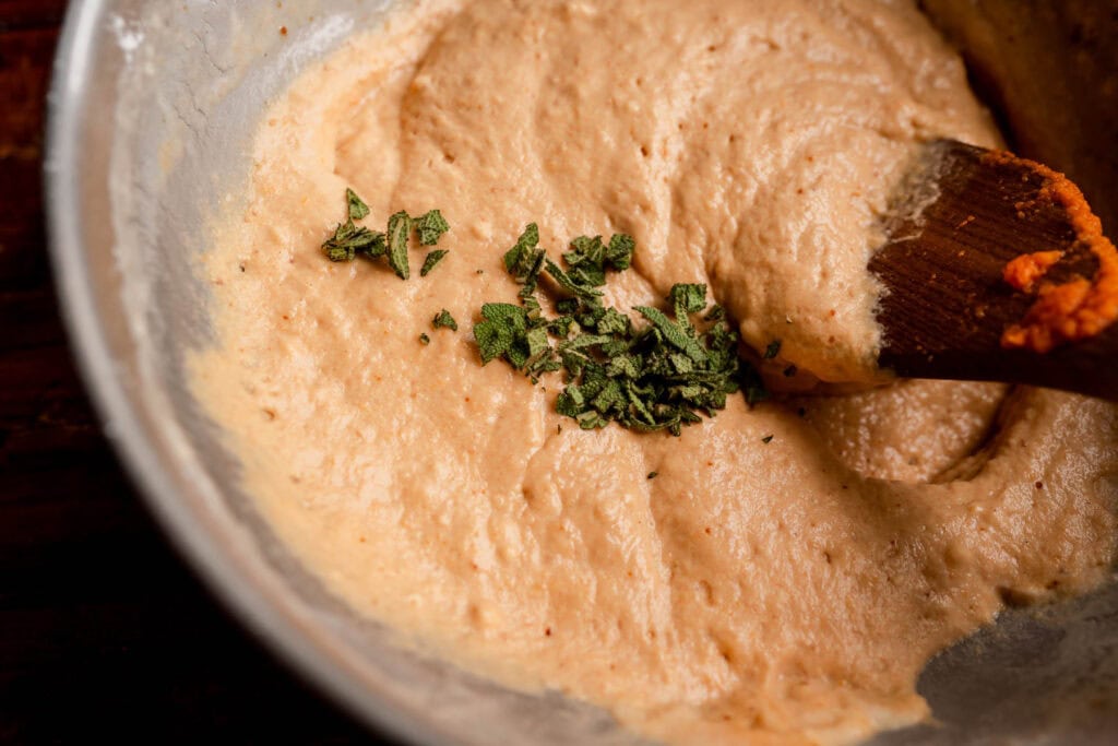 A wooden spoon stirs pumpkin butter swim biscuit batter sprinkled with chopped fresh sage in a metal mixing bowl.