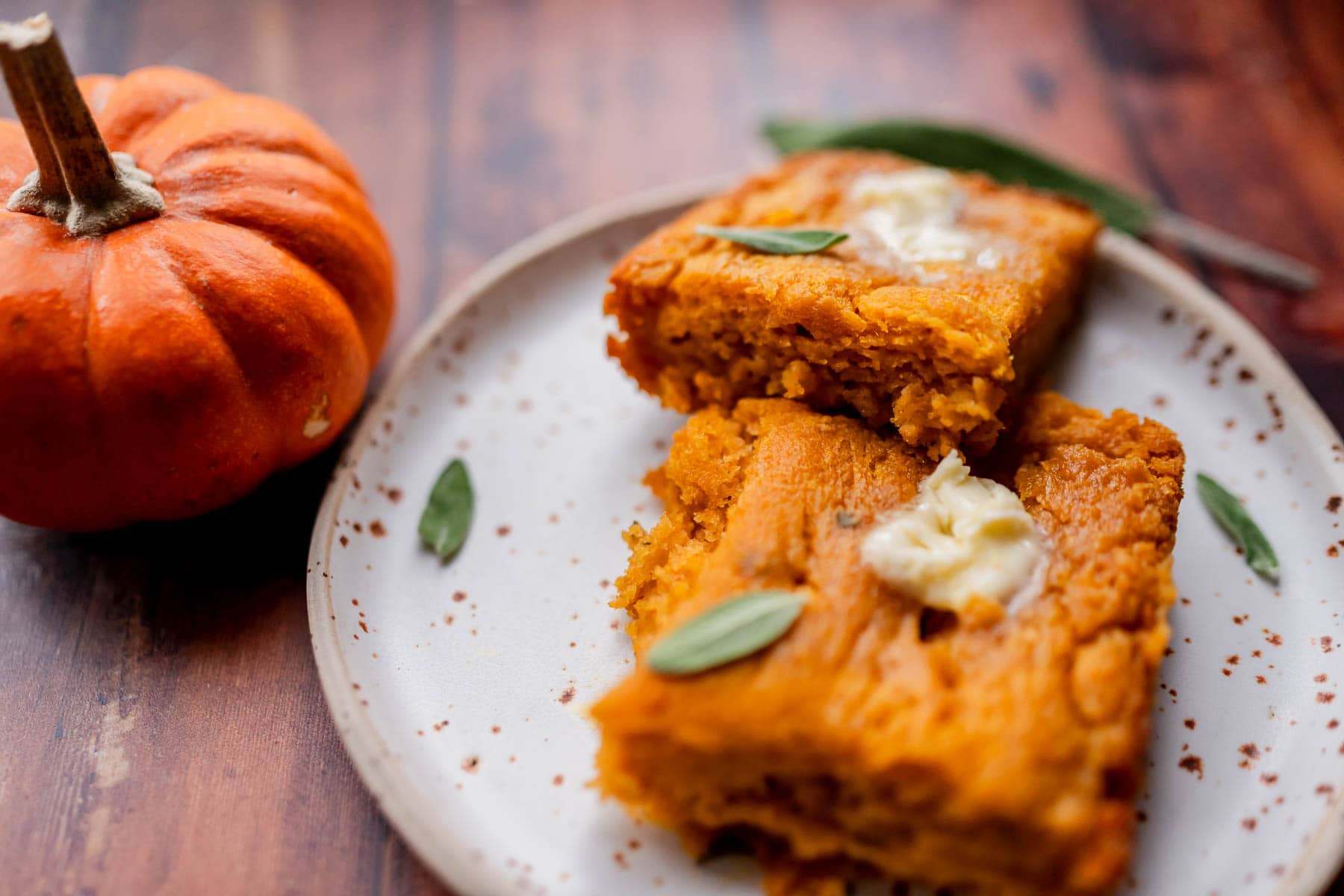 Two squares of pumpkin butter swim biscuits topped with butter and sage on a speckled plate next to a small pumpkin.