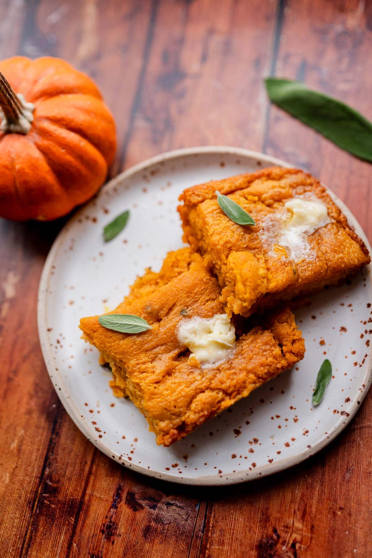 Two squares of pumpkin butter swim biscuits topped with butter and sage leaves on a plate next to a small pumpkin.
