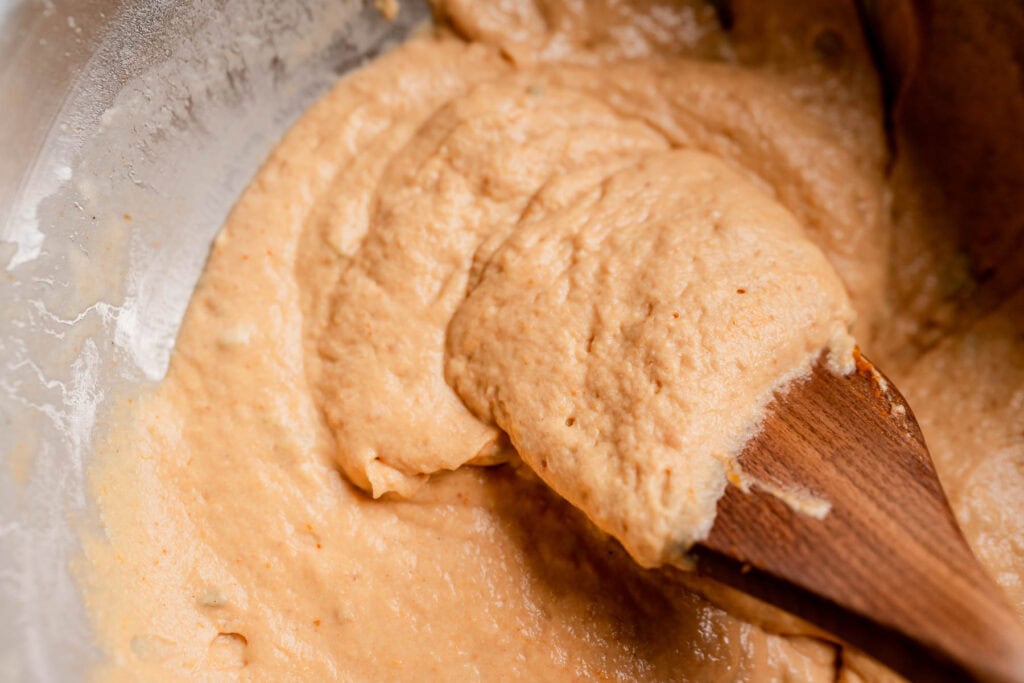 A wooden spoon mixing light orange batter for pumpkin butter swim biscuits in a metal bowl.