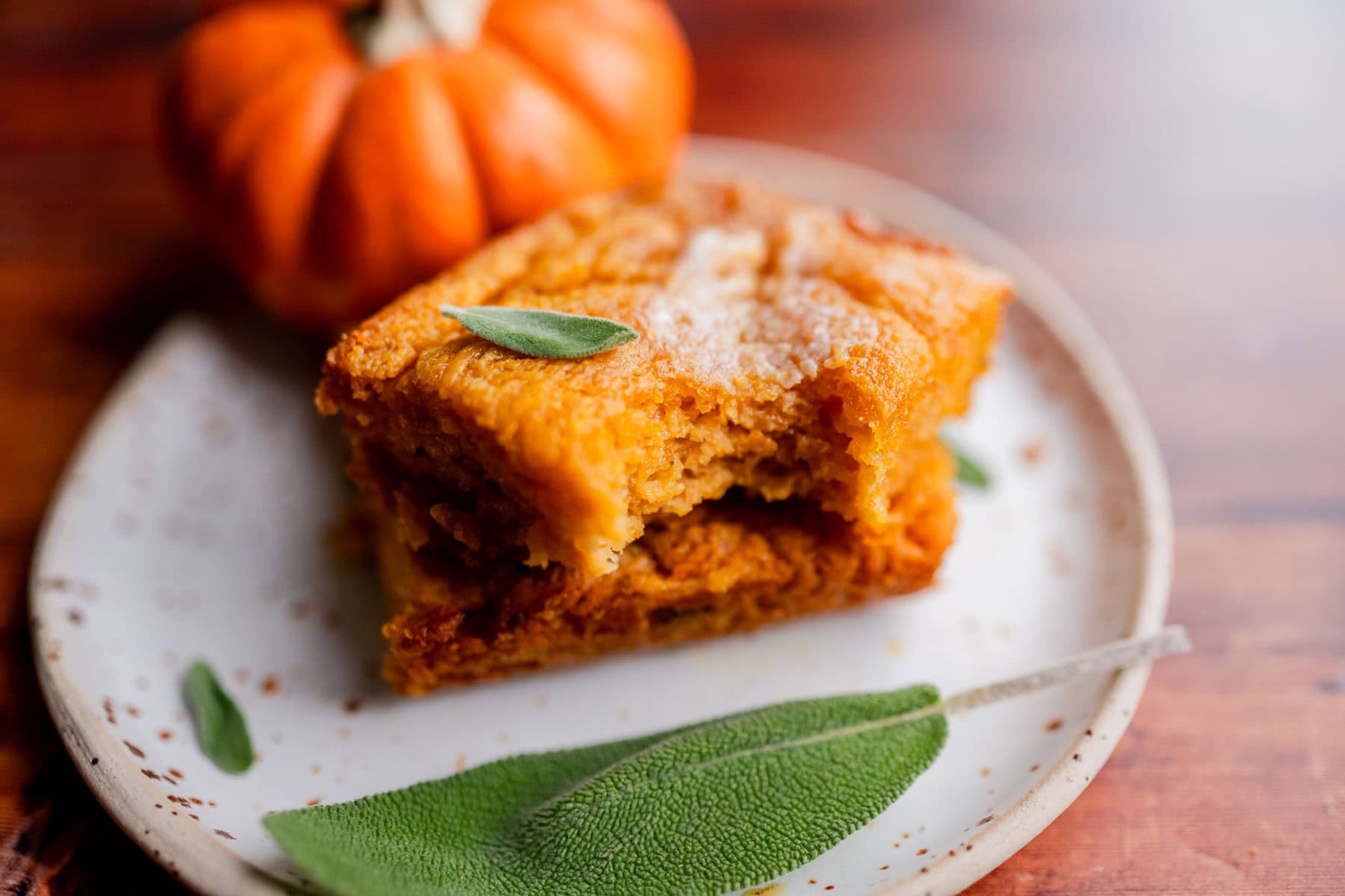 A piece of pumpkin butter swim biscuit with a bite taken out, garnished with sage leaves on a speckled plate next to a small pumpkin.