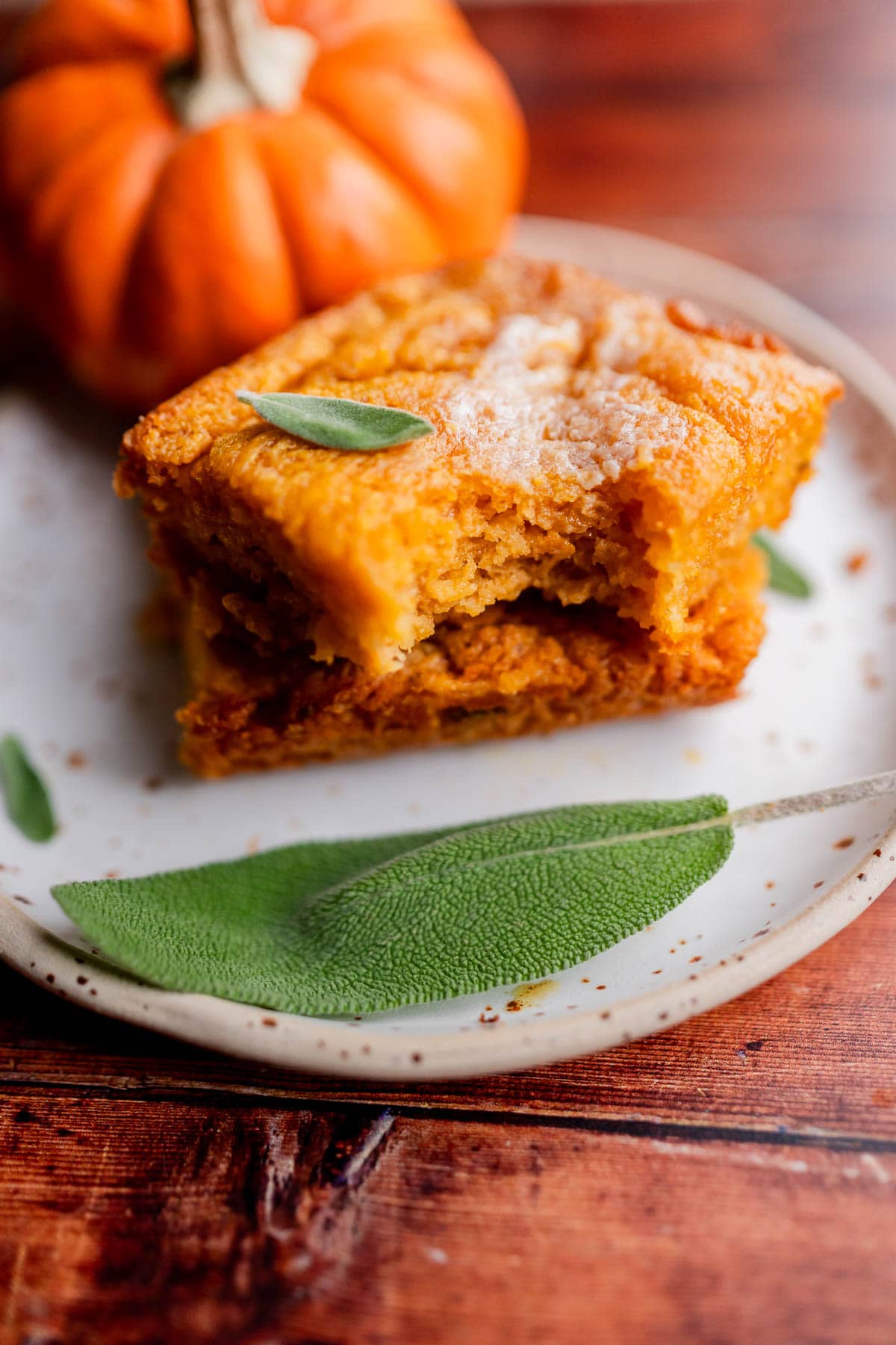 Two stacked squares of pumpkin butter swim biscuits, one with a bite taken out, garnished with fresh sage leaves on a plate next to a small pumpkin.